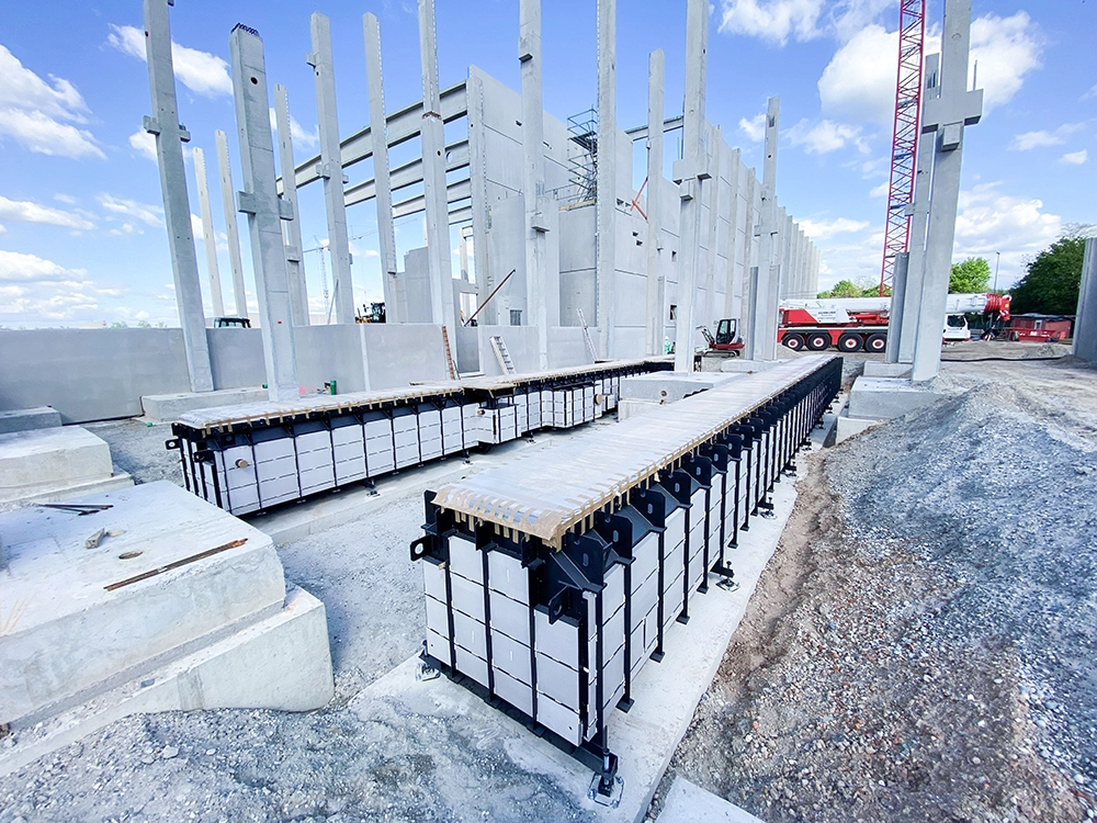 Montage einer großformatigen truckcon Stahlfertiggrube auf einer Baustelle: Zwei Kräne heben die vormontierte Werkstattgrube vor einer modernen Industriehalle in Position. Die Aufnahme zeigt die präzise Installation schwerer Werkstatttechnik für Nutzfahrzeugwerkstätten.